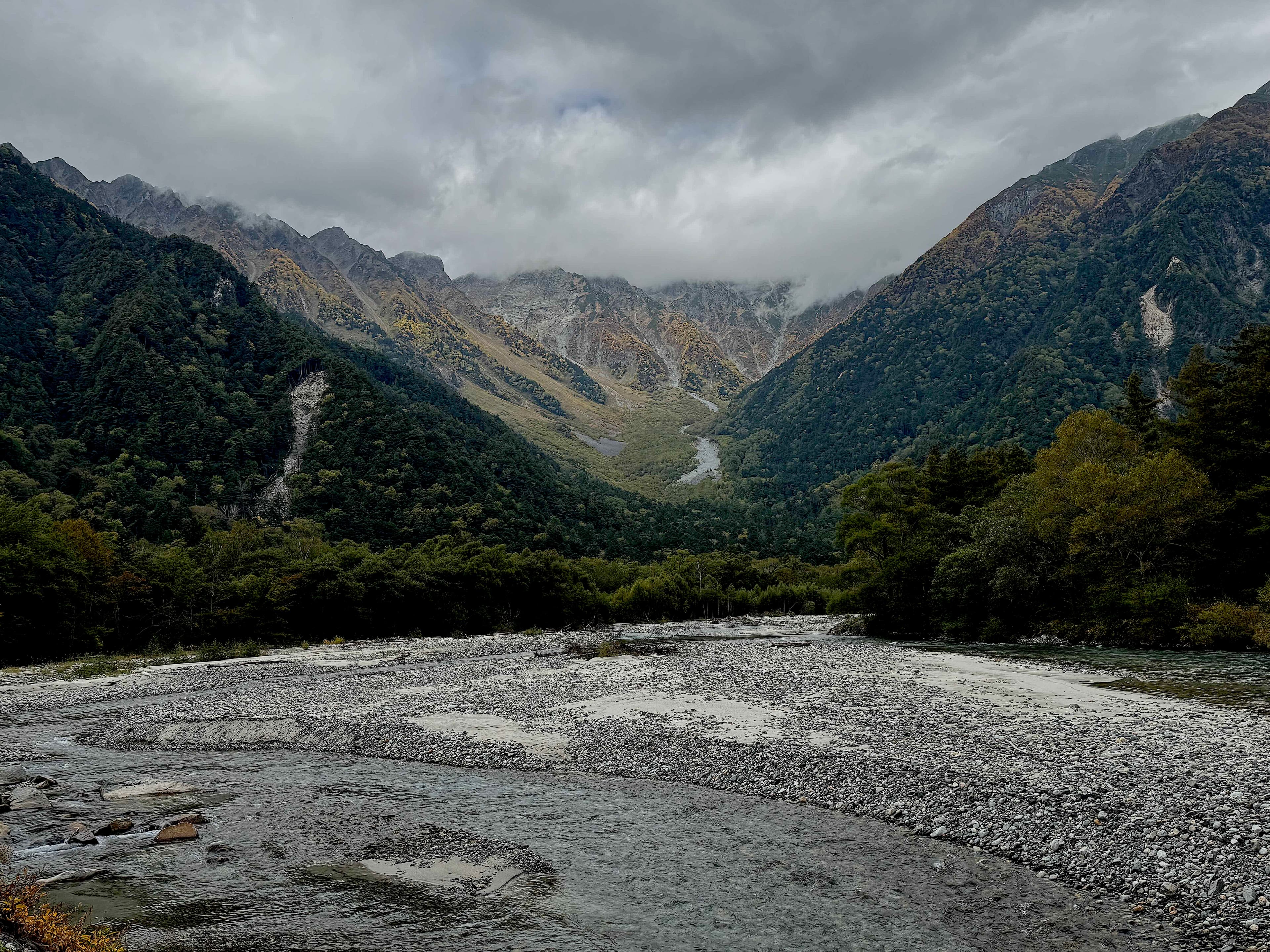 Wide view of a river flowing through a rocky valley with dense green and autumn-colored forests, framed by cloud-covered mountains